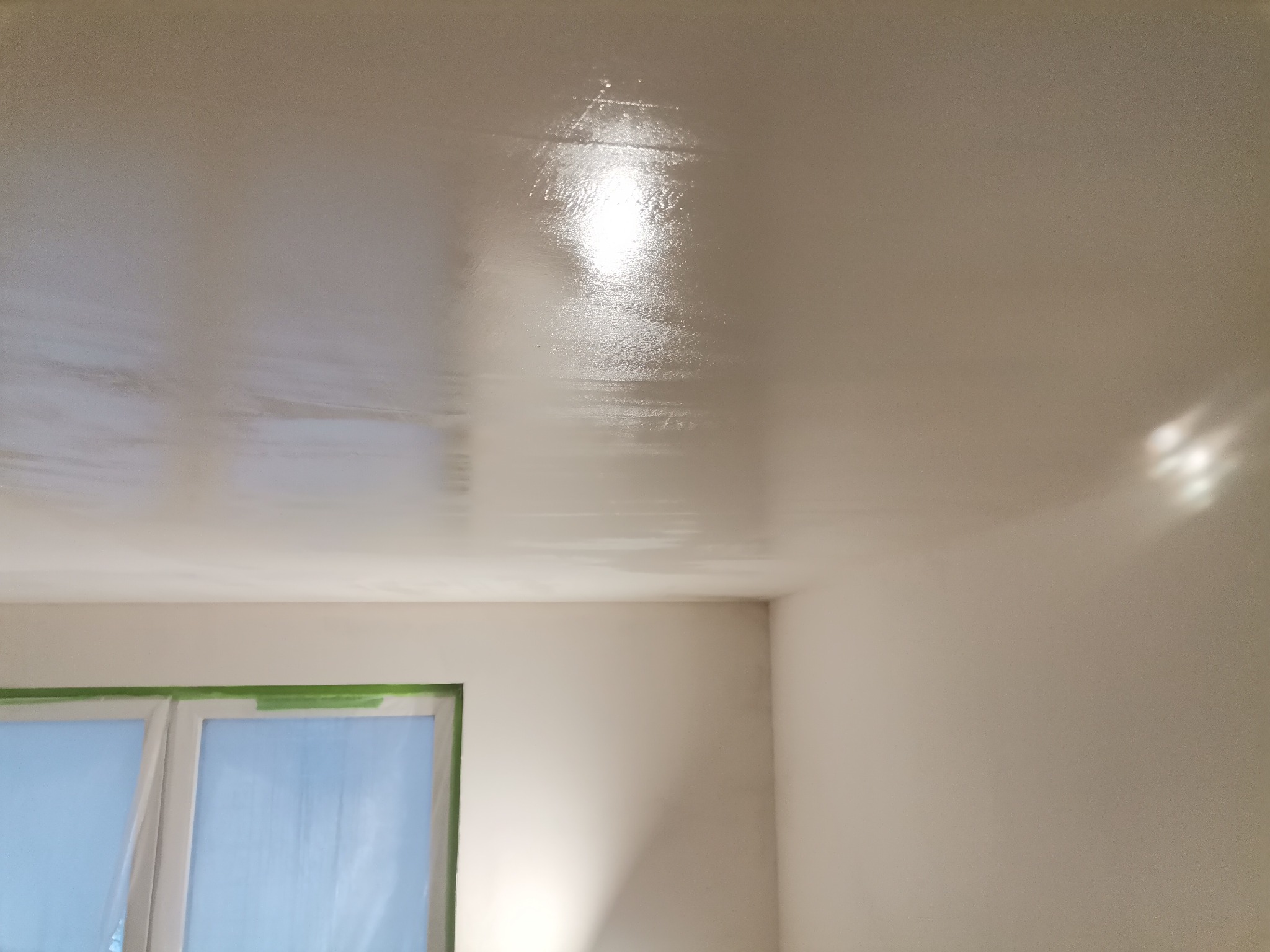 Świeżo pomalowany sufit i ściana w mieszkaniu w Warszawie, z widocznym odbiciem światła i zabezpieczonym oknem taśmą malarską.