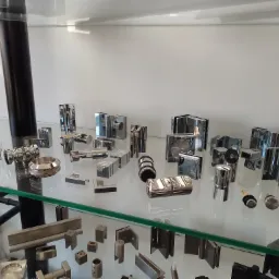 Ekspozycja różnorodnych okuć i akcesoriów do szkła, w tym zawiasów, uchwytów i mocowań, prezentowana na szklanej półce w jasnym wnętrzu.