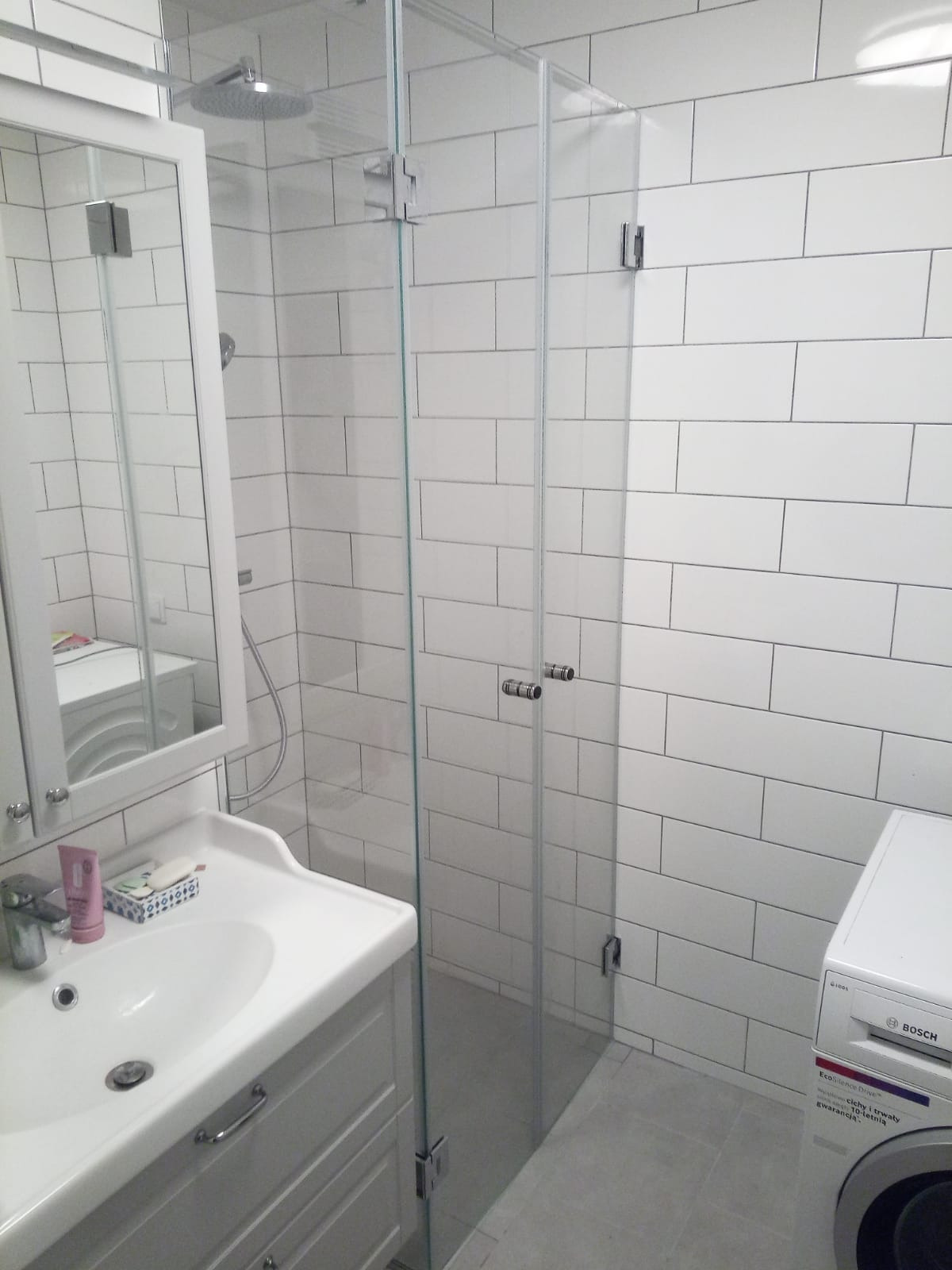 Szklana kabina prysznicowa z chromowanymi zawiasami i uchwytami w łazience z białymi, prostokątnymi płytkami ułożonymi w cegiełkę, widoczna umywalka z szafką i pralka Bosch.