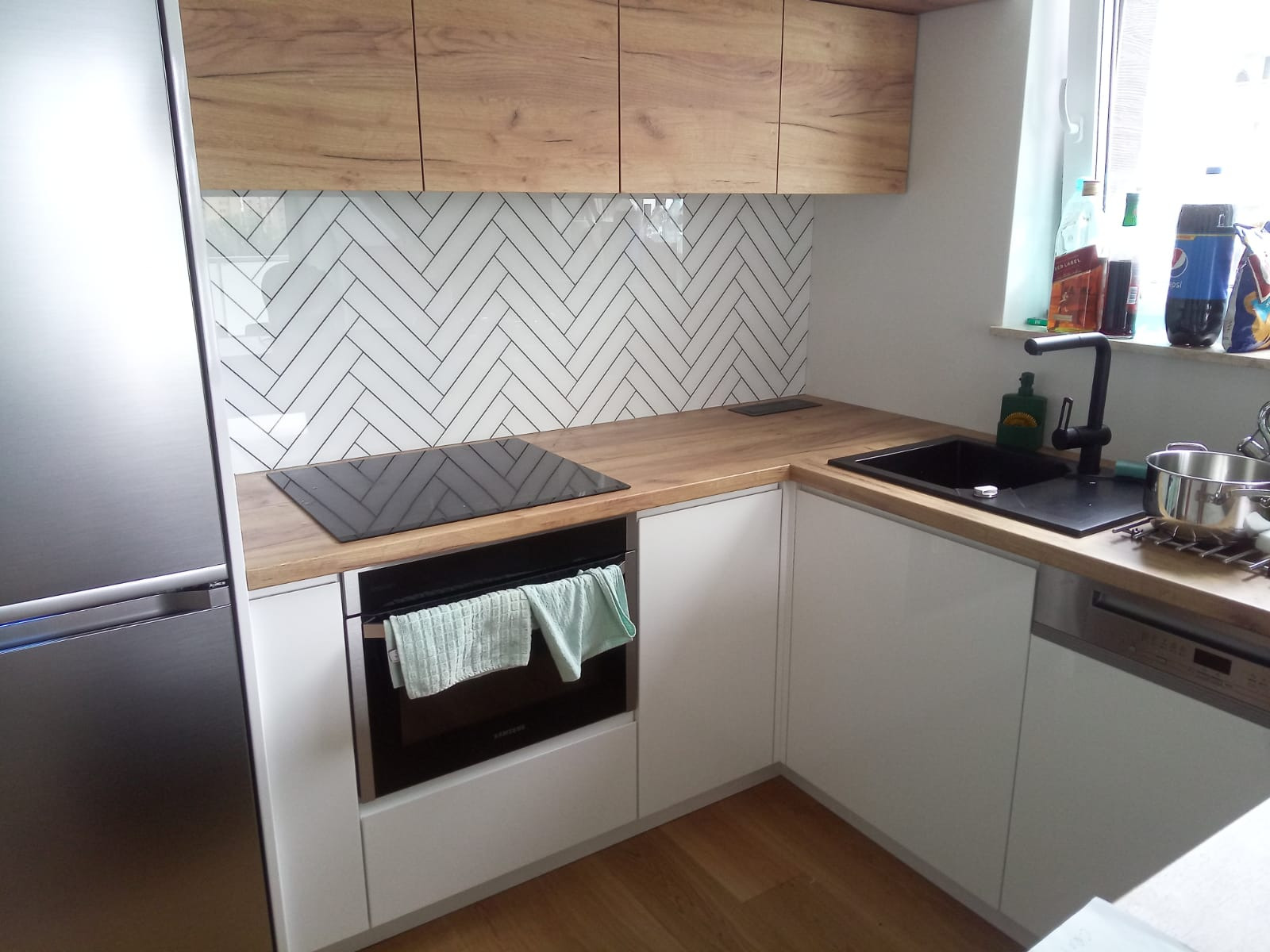 Nowoczesna kuchnia z białymi szafkami, drewnianym blatem i szklanym panelem nad blatem ułożonym w jodełkę, czarna płyta indukcyjna i zlewozmywak, ręczniki na piekarniku.