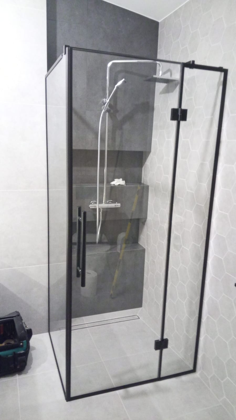 Nowoczesna kabina prysznicowa z czarnymi profilami, szklana, z deszczownicą i dodatkową słuchawką, umieszczona w łazience z szarą armaturą i mozaiką na ścianie, widoczna skrzynka z narzędziami.