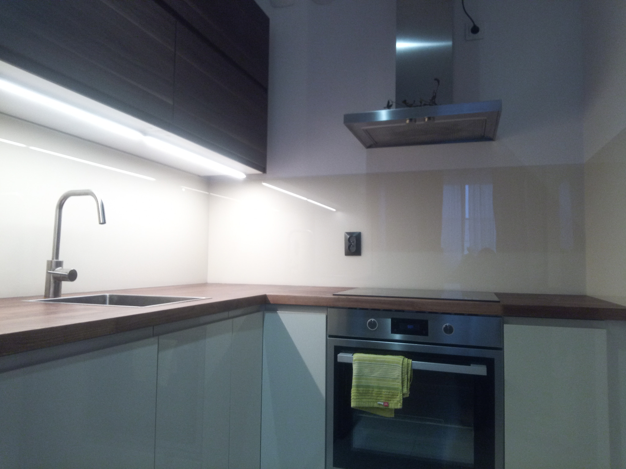 Nowoczesna kuchnia z jasnymi szafkami, drewnianym blatem, szklanym panelem ściennym i oświetleniem LED pod szafkami wiszącymi. Na piekarniku wisi zielona ściereczka.