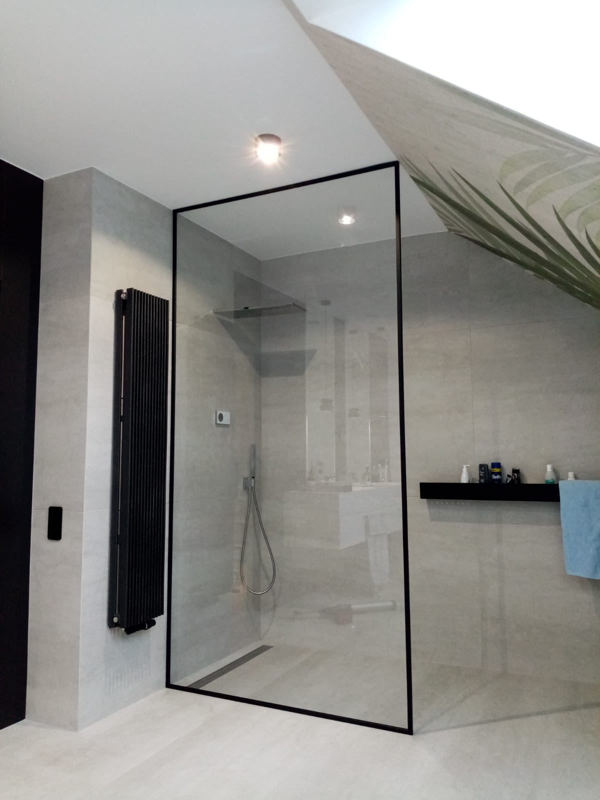 Nowoczesna kabina prysznicowa walk-in z czarną ramą, zamontowana w łazience z szarymi płytkami, widoczny czarny grzejnik drabinkowy i półka z kosmetykami, fragment dekoracji sufitowej z motywem liści.