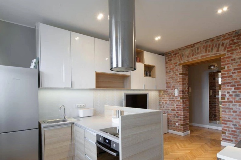 Nowoczesna kuchnia z wyspą, białymi szafkami na wysoki połysk i ceglaną ścianą w tle, oświetlenie punktowe.