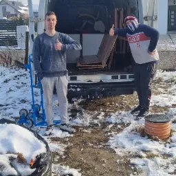 Dwóch mężczyzn przed otwartym busem załadowanym meblami, jeden z wózkiem transportowym, obaj pokazują kciuk w górę, teren pokryty śniegiem.