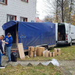 Dwóch mężczyzn wyładowuje białą pralkę z niebieskiej przyczepy transportowej przed budynkiem mieszkalnym, obok kartony i zapakowane meble.