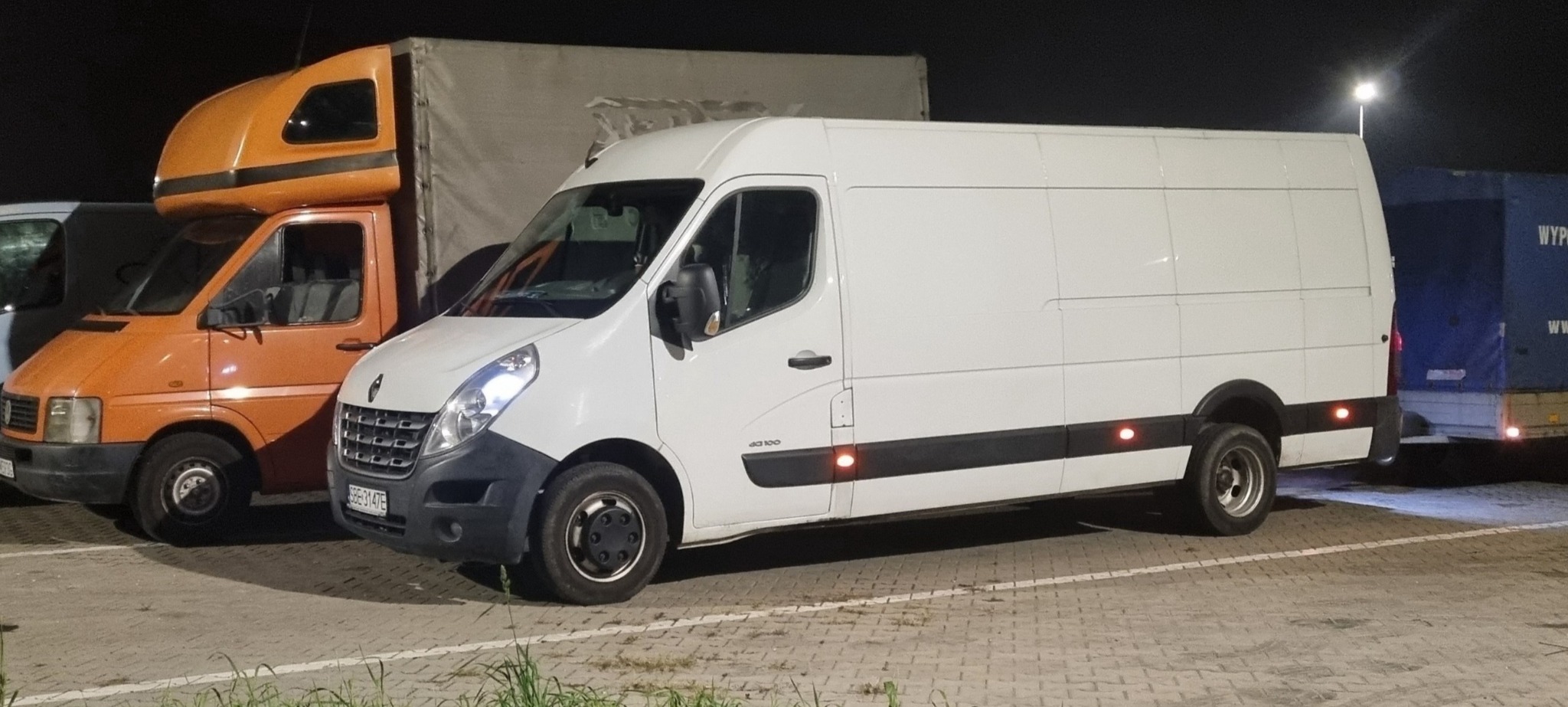 Biały samochód dostawczy Renault Master zaparkowany na parkingu, obok pomarańczowego i niebieskiego pojazdu ciężarowego z naczepą, nocne oświetlenie