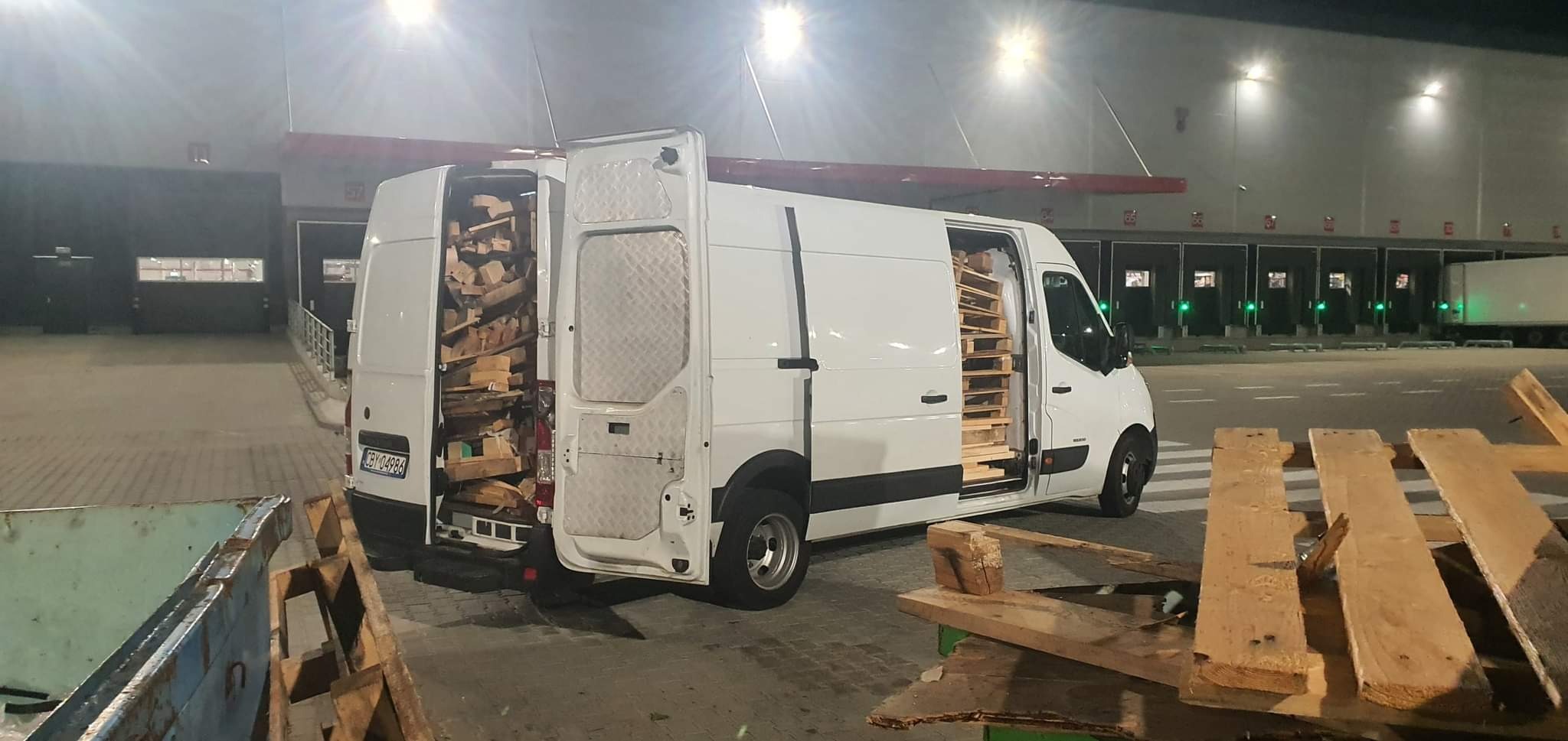 Biały furgon z otwartymi drzwiami, wypełniony drewnianymi paletami, zaparkowany przy rampie załadunkowej magazynu.