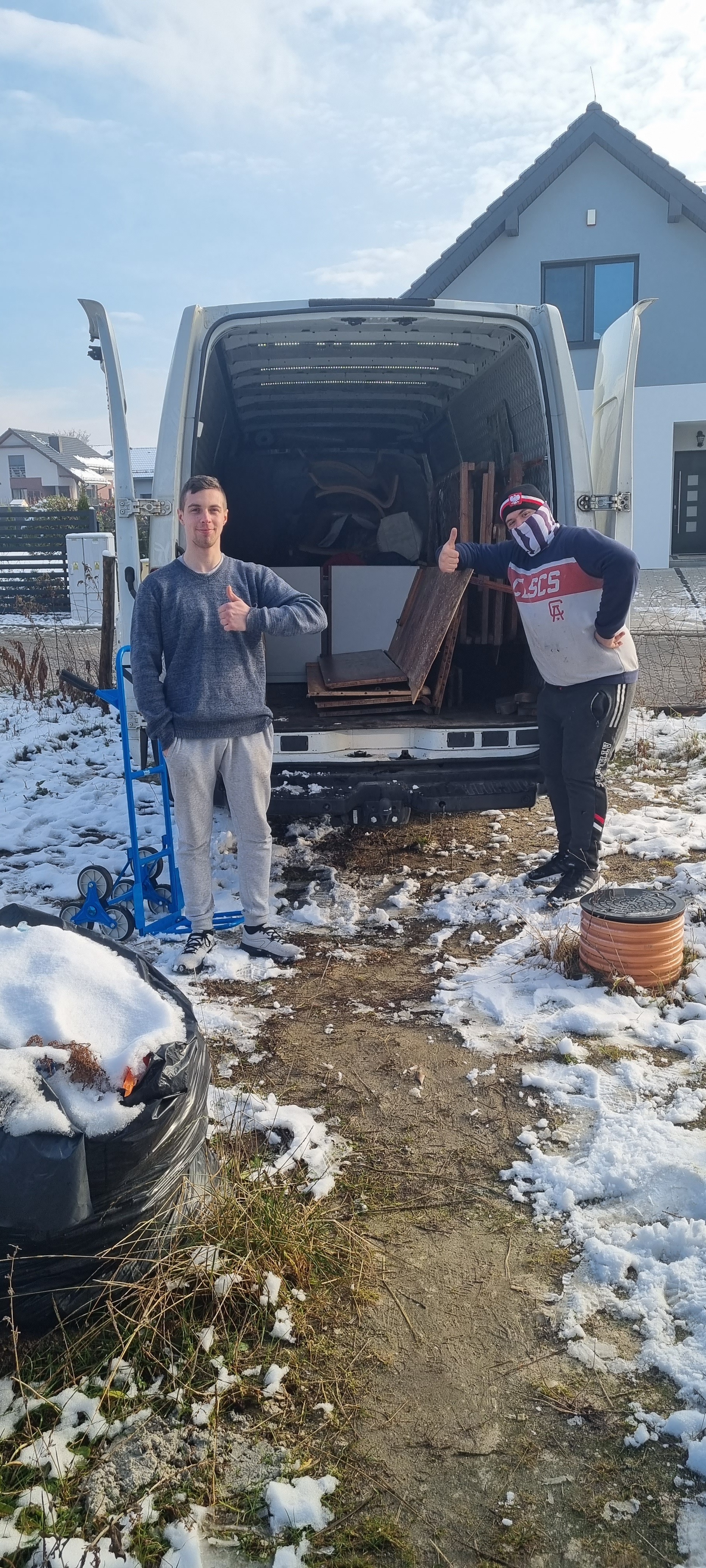 Dwóch mężczyzn przed otwartym busem załadowanym meblami, jeden z wózkiem transportowym, obaj pokazują kciuk w górę, teren pokryty śniegiem.