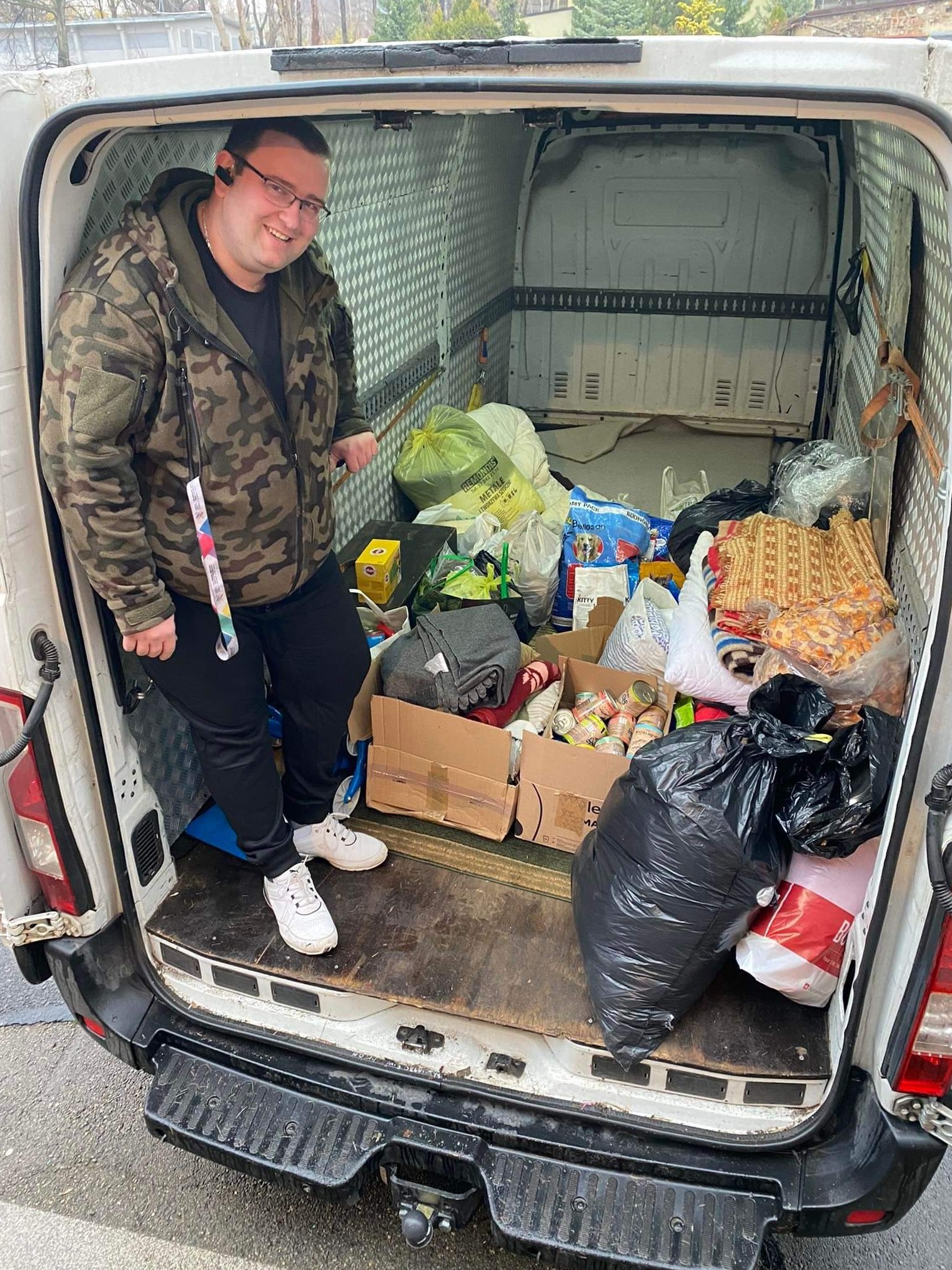 Uśmiechnięty mężczyzna w kurtce moro stoi w otwartym bagażniku białego vana wypełnionego kartonami, workami i różnymi przedmiotami, przygotowanymi do transportu.