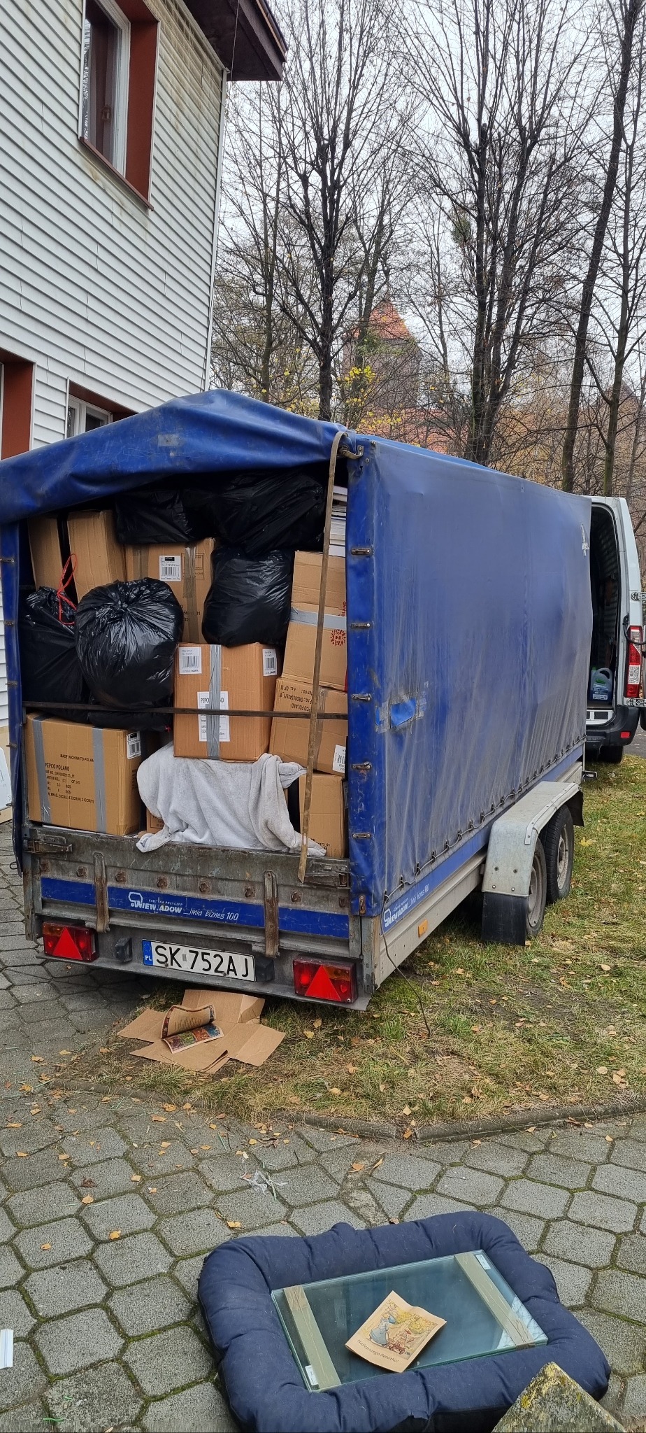 Przyczepa wypełniona kartonami i czarnymi workami, częściowo przykryta niebieską plandeką, zaparkowana na kostce brukowej przed domem. Na ziemi leżą rozpakowane kartony i poduszka z szybą.