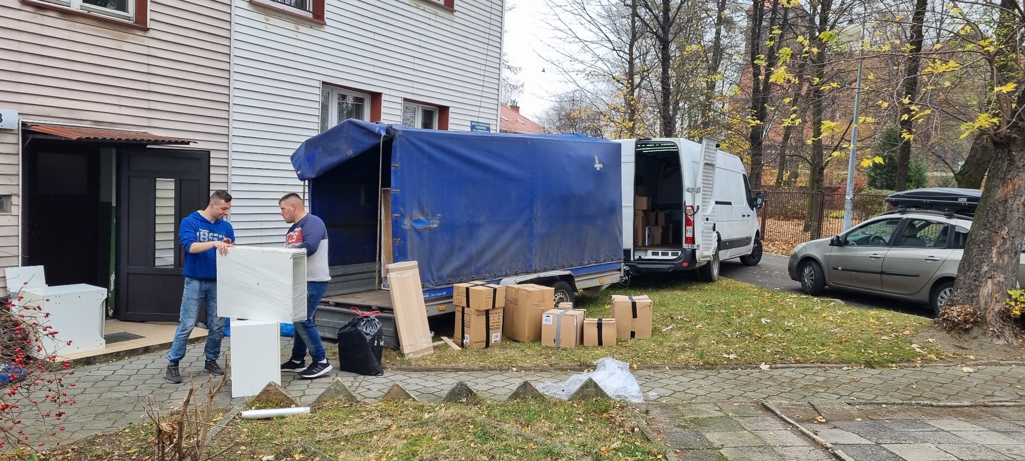 Dwóch mężczyzn przenosi owinięte folią białe szafki z przyczepy z niebieską plandeką na tle budynku z drewnianą elewacją i zaparkowanego szarego samochodu z bagażnikiem dachowym.
