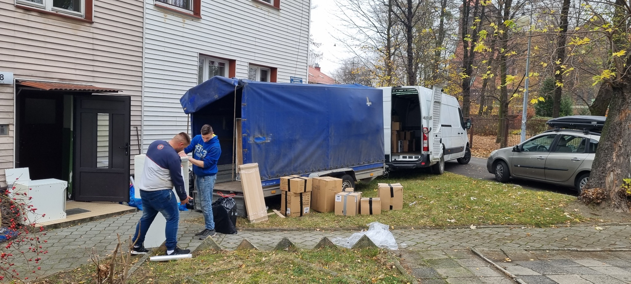 Dwóch mężczyzn wyładowuje białą pralkę z niebieskiej przyczepy transportowej przed budynkiem mieszkalnym, obok kartony i zapakowane meble.