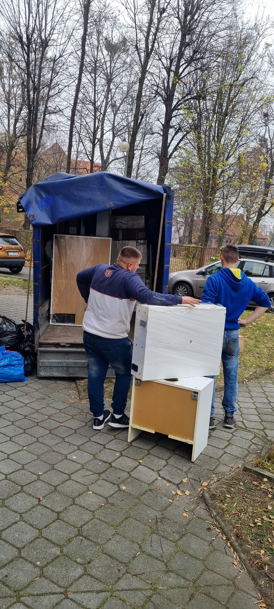 Dwóch mężczyzn wyładowuje z samochodu dostawczego meble, w tym białą szafkę owiniętą folią stretch, na brukowanym chodniku w jesienny dzień.