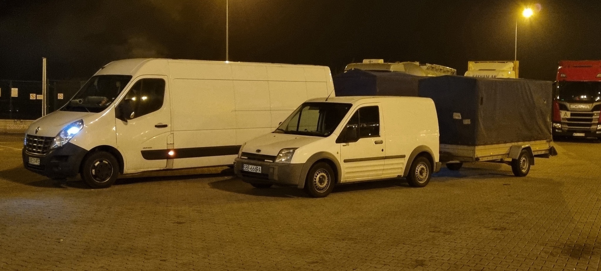 Dwa białe busy i przyczepka z plandeką stojące na parkingu nocą, w tle widoczna ciężarówka Scania.