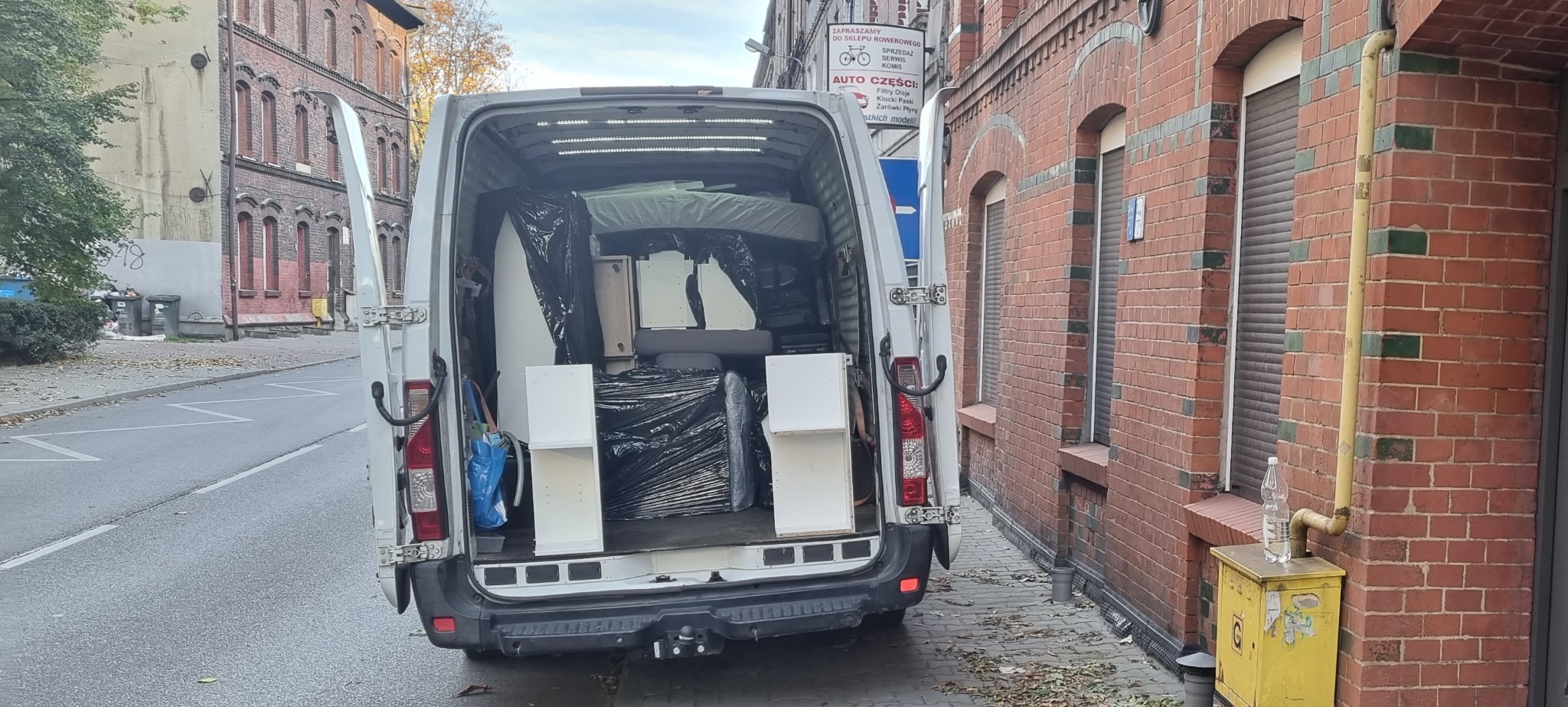 Otwarty tył białego busa wypełnionego meblami, niektóre owinięte w czarną folię, na tle ceglanego budynku z zamkniętymi roletami i żółtą skrzynką gazową.