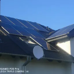 Panele fotowoltaiczne zamontowane na dachu domu jednorodzinnego z ceglanym kominem i anteną satelitarną, błękitne niebo w tle.