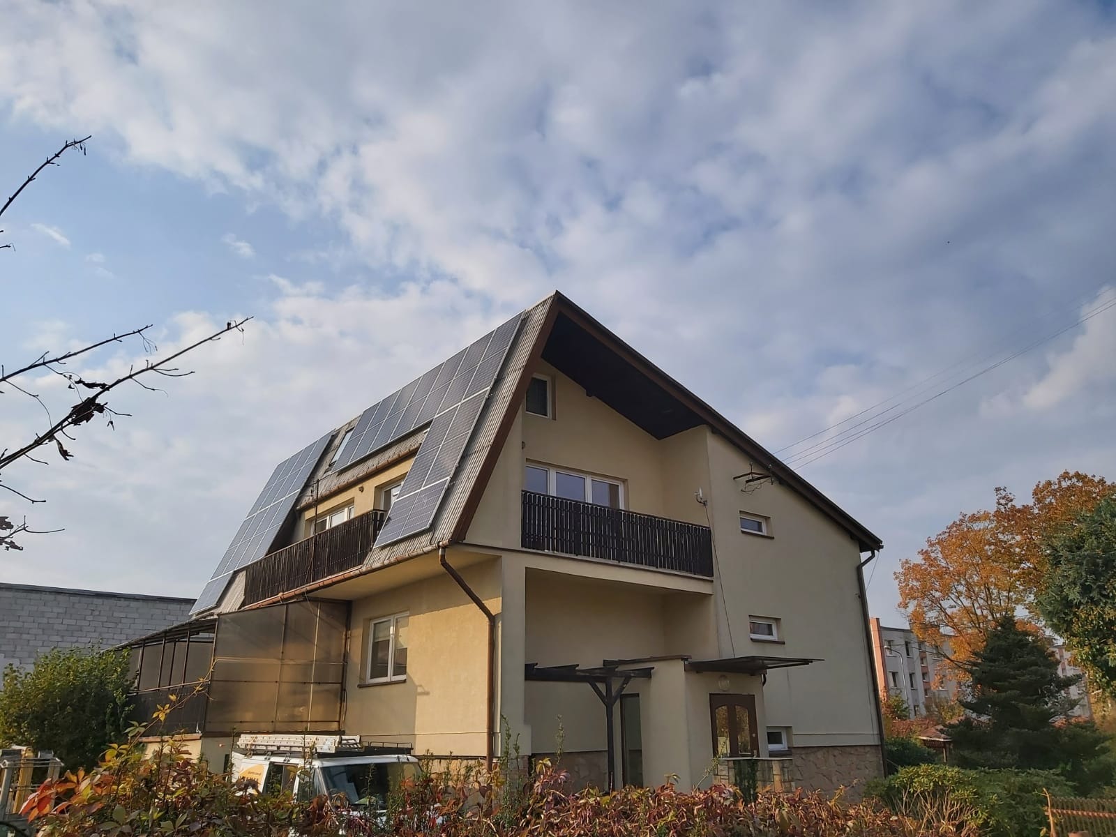 Dwupiętrowy dom jednorodzinny z panelami słonecznymi na dachu, widoczne balkony z ciemnymi balustradami i częściowo osłonięty taras na parterze, na pierwszym planie krzewy z brązowymi liśćmi.