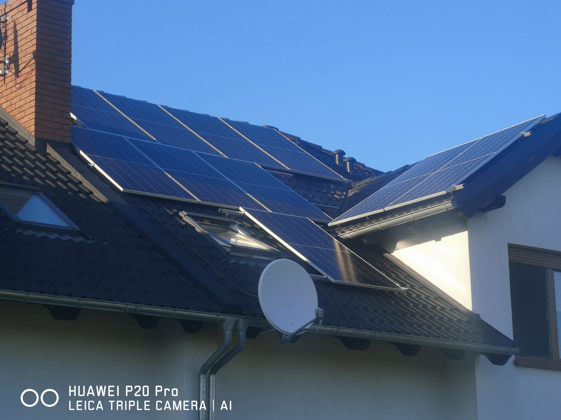 Panele fotowoltaiczne zamontowane na dachu domu jednorodzinnego z ceglanym kominem i anteną satelitarną, błękitne niebo w tle.