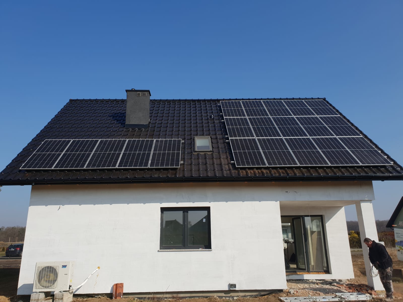 Biały dom jednorodzinny z czarnym dachem pokrytym panelami fotowoltaicznymi w słoneczny dzień.