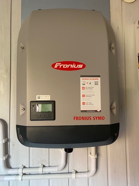 Szary inwerter Fronius Symo zamontowany na drewnianej ścianie z widocznymi białymi rurami instalacyjnymi poniżej.