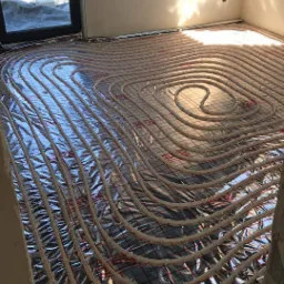 Białe rurki układane w spiralny wzór na folii odblaskowej z czerwoną siatką, przygotowane do zalania wylewką pod ogrzewanie podłogowe, widok z góry w surowym wnętrzu z oknem.