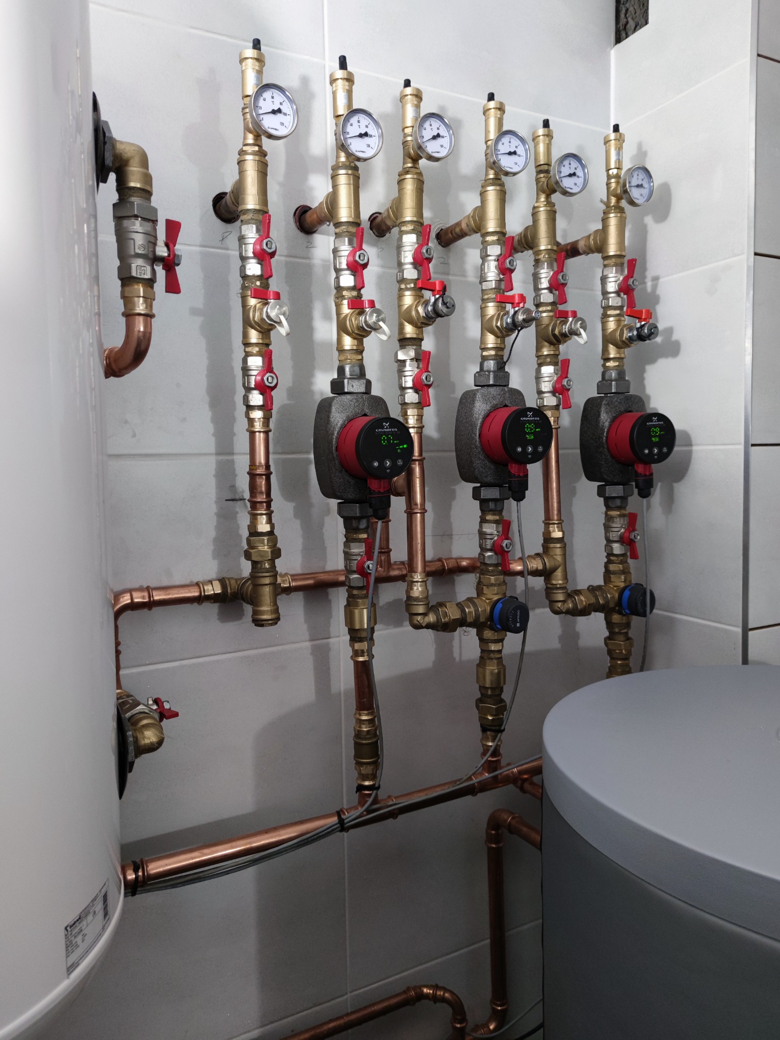 Rozbudowany system rur miedzianych z zaworami, manometrami i pompami obiegowymi marki Grundfos, zamontowany na ścianie z płytek ceramicznych.