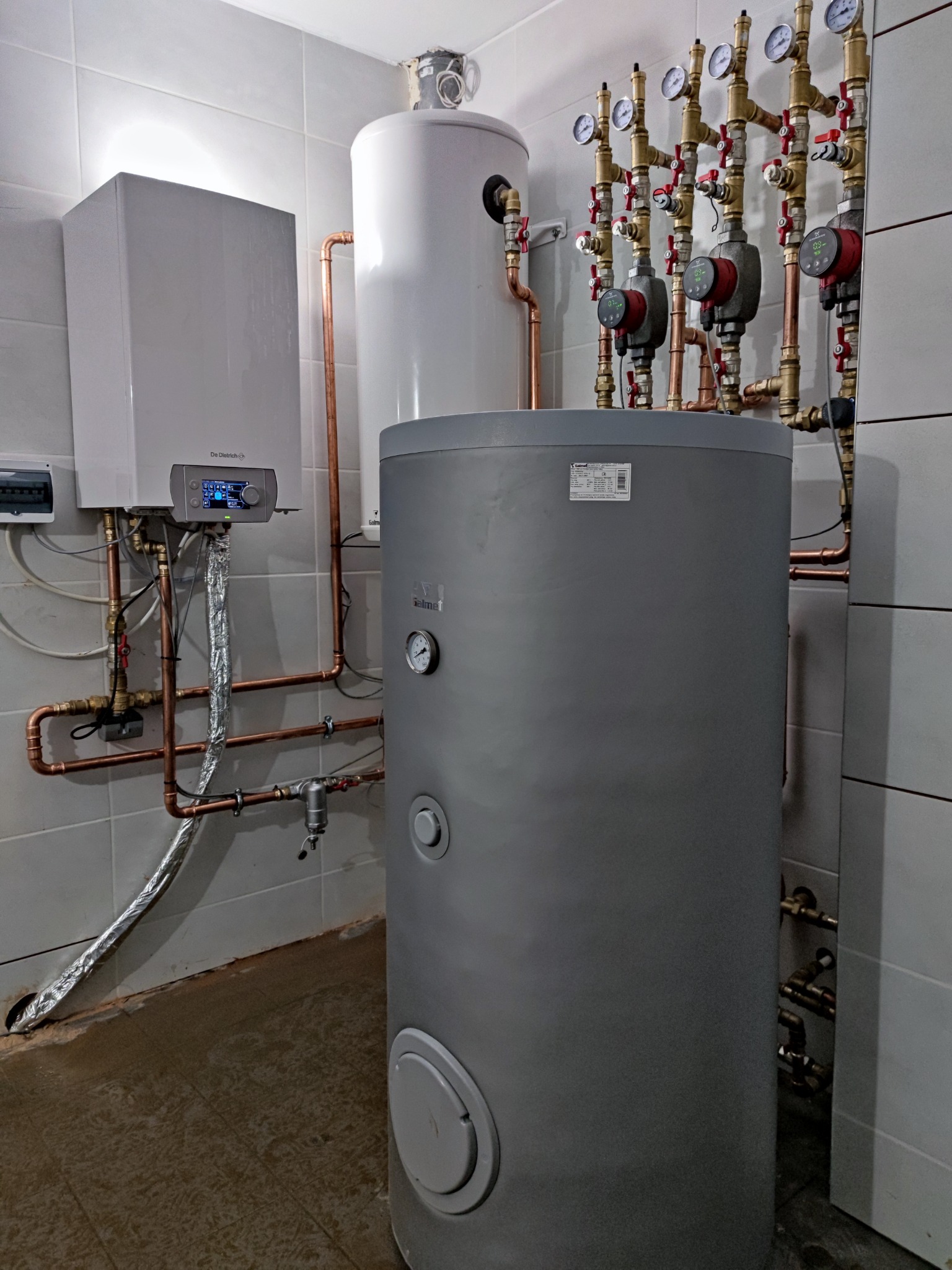Kompaktowa kotłownia w pomieszczeniu z szarymi płytkami, z widocznym kotłem gazowym De Dietrich, zasobnikiem ciepłej wody, grupą pompową z manometrami i miedzianymi rurami.