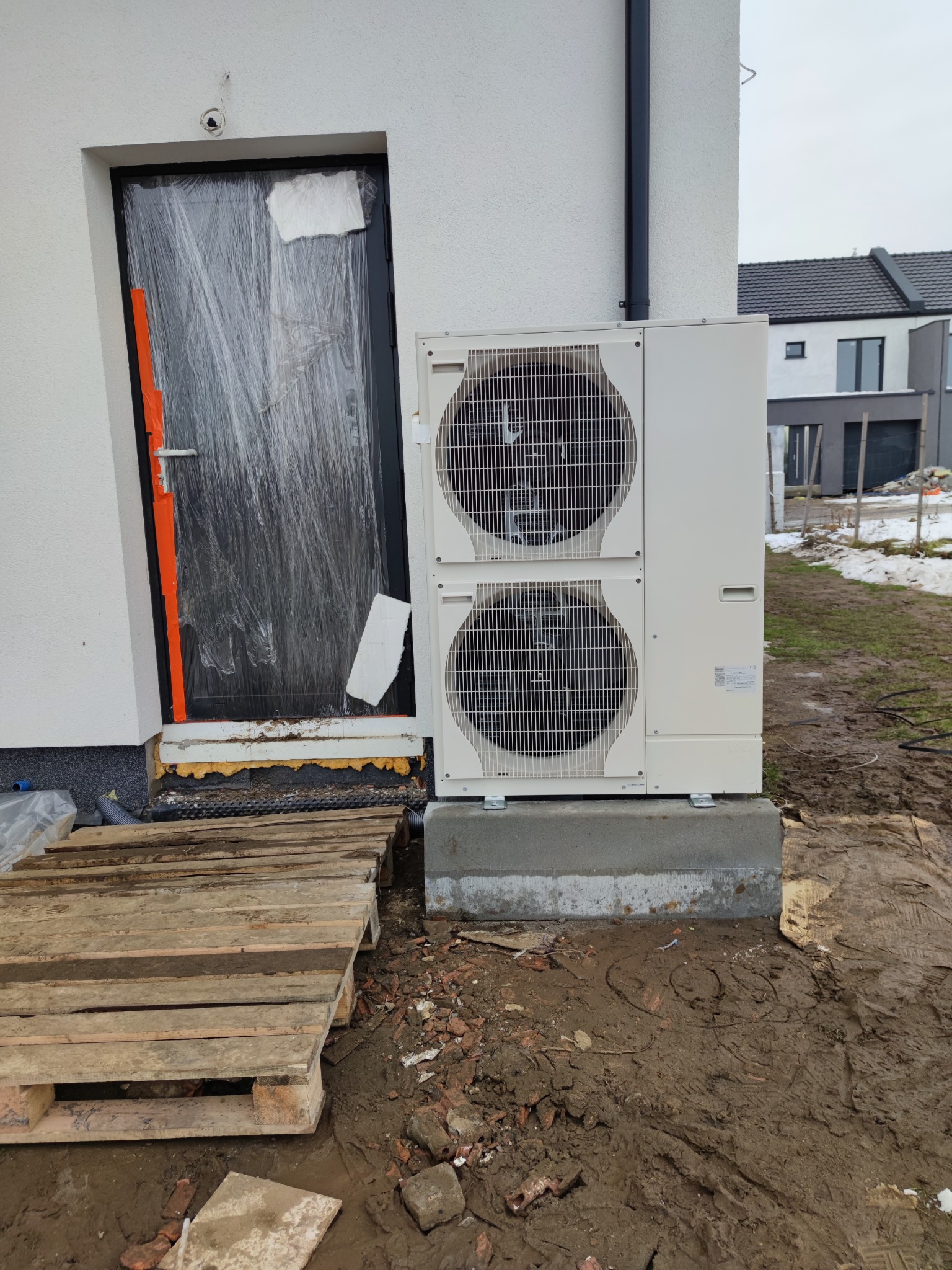Biała pompa ciepła zamontowana na betonowym fundamencie przy ścianie budynku w trakcie instalacji; drzwi wejściowe zabezpieczone folią ochronną; teren wokół rozkopany i błotnisty.