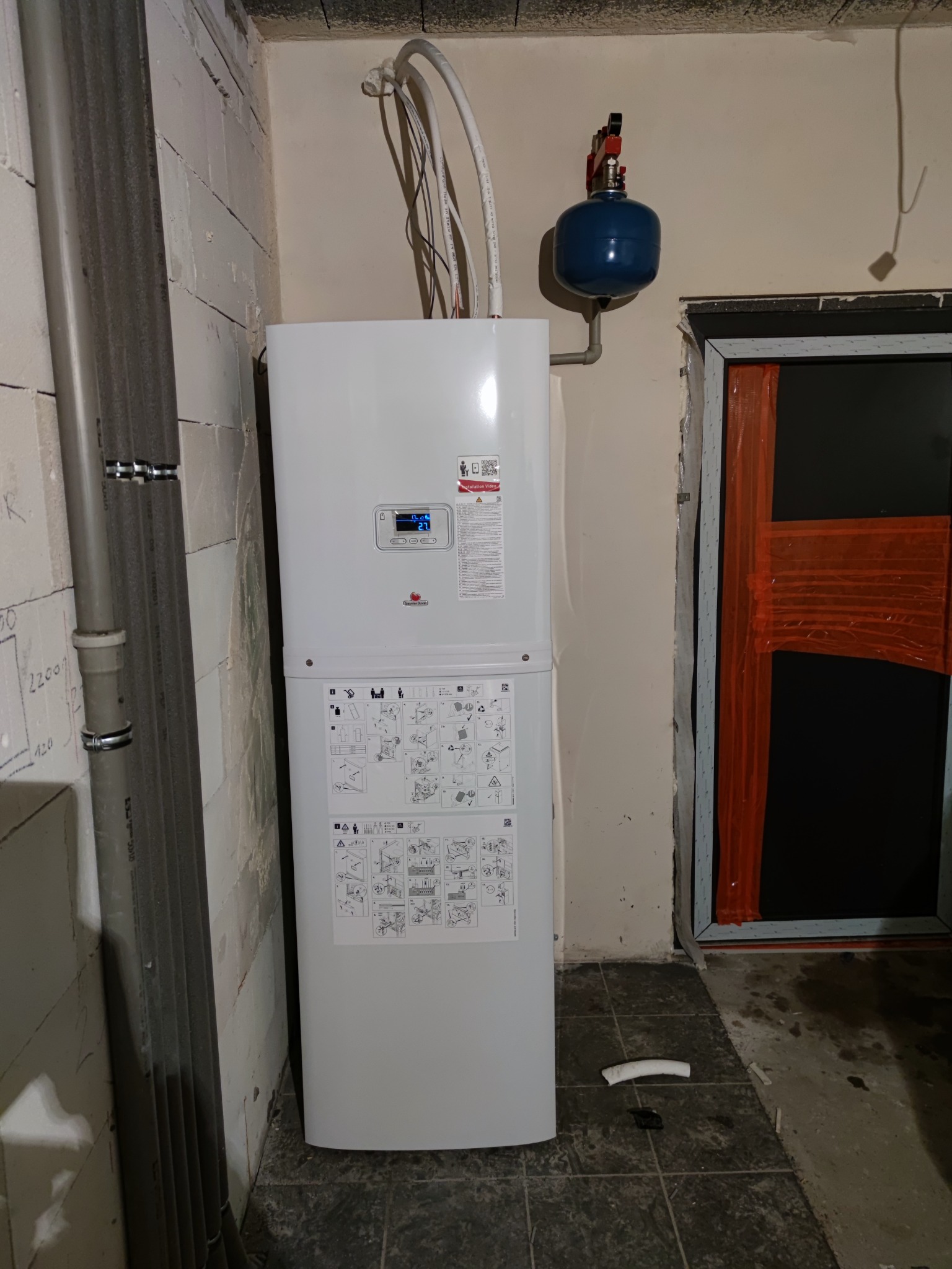 Biały piec centralnego ogrzewania z niebieskim zbiornikiem wyrównawczym zamontowany w surowym pomieszczeniu z betonowych pustaków i ciemną podłogą.