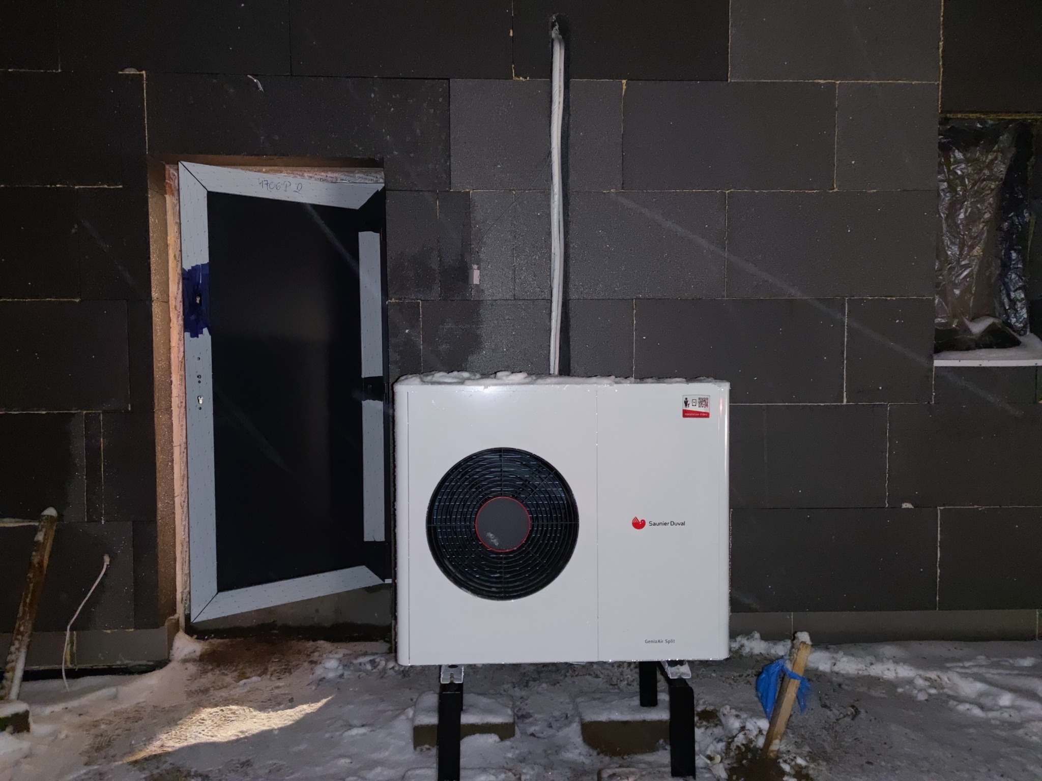 Biała pompa ciepła Saunier Duval GeniaAir Split zamontowana na zewnątrz budynku z ciemnej cegły w zimowej scenerii, widoczne wystające przewody i fragment okna zabezpieczonego folią.