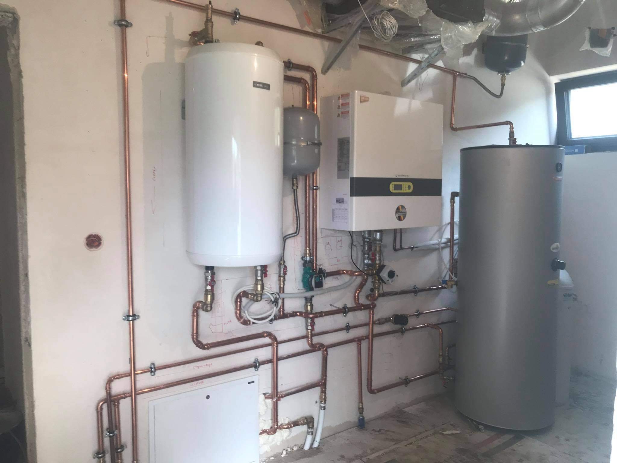 Kompletna instalacja grzewcza w trakcie montażu: biały zasobnik ciepłej wody, piec gazowy z wyświetlaczem, szary zbiornik wyrównawczy i drugi zasobnik, połączone miedzianymi rurami w surowym...