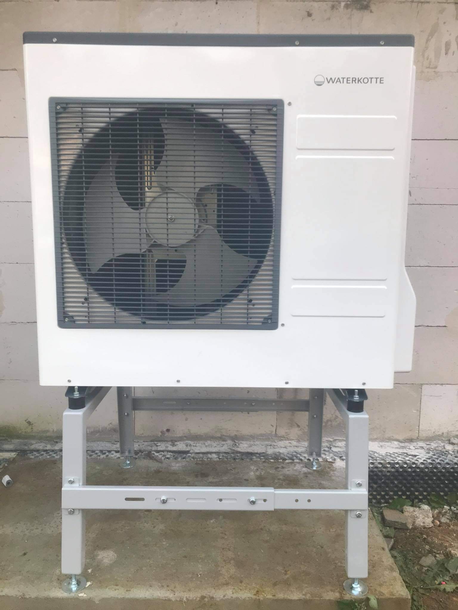 Biała jednostka zewnętrzna pompy ciepła marki Waterkotte, zamontowana na metalowym stelażu z regulowanymi nóżkami, ustawiona na betonowym podłożu przed ścianą z bloczków betonowych.