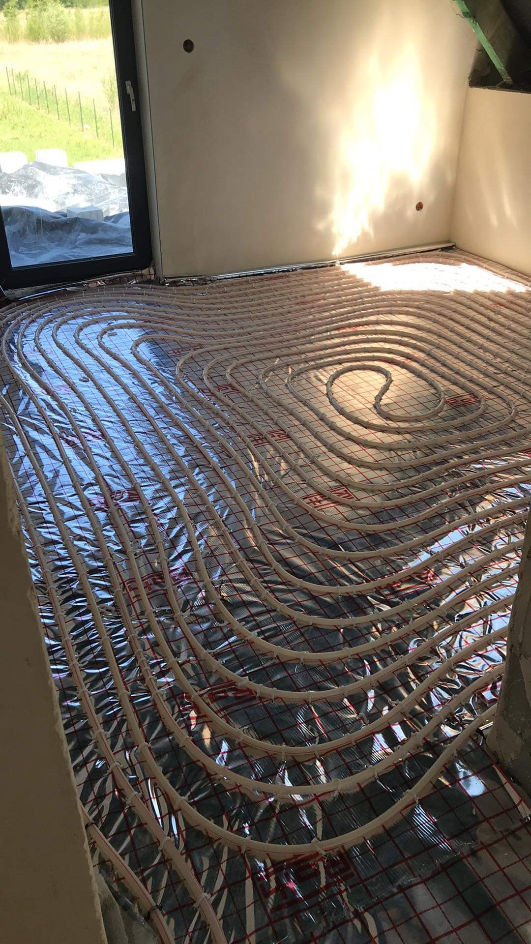 Białe rurki układane w spiralny wzór na folii odblaskowej z czerwoną siatką, przygotowane do zalania wylewką pod ogrzewanie podłogowe, widok z góry w surowym wnętrzu z oknem.