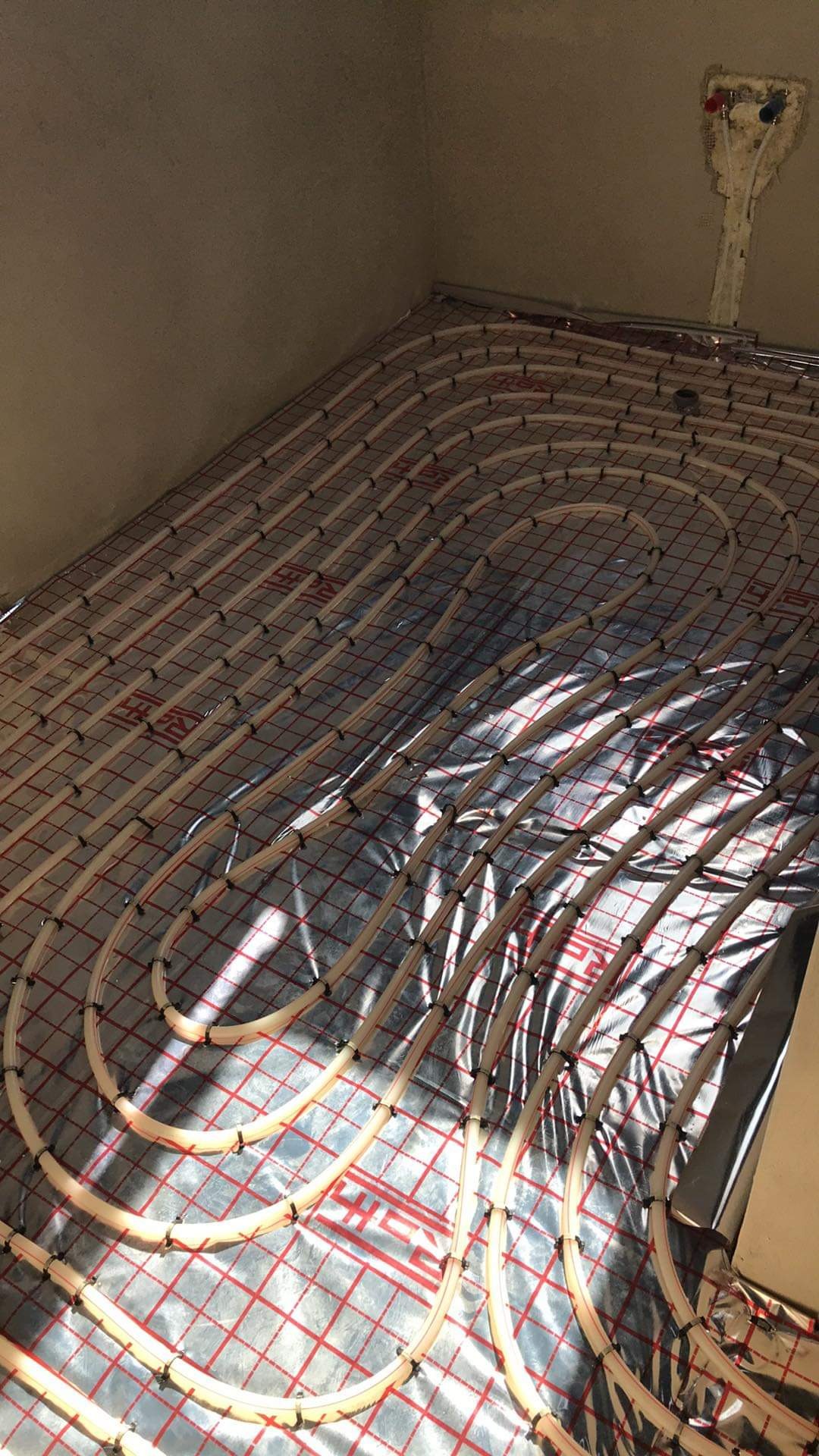 Rury ogrzewania podłogowego ułożone na srebrnej folii z czerwoną siatką, widoczne przyłącza do instalacji na ścianie w surowym stanie.