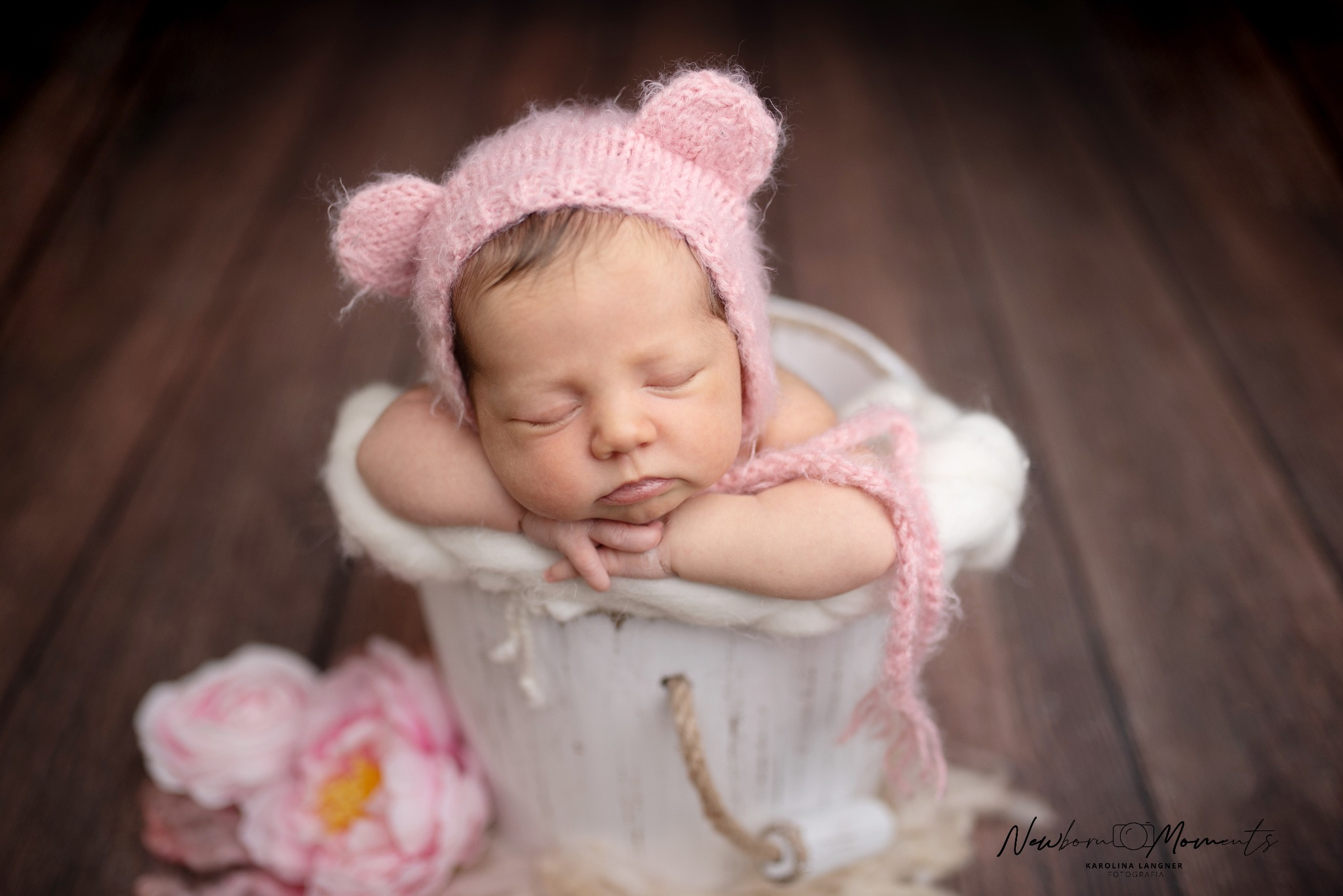 Śpiący noworodek w różowej czapce z uszkami, ułożony w białym wiaderku z futerkiem, obok różowe piwonie na drewnianym tle.