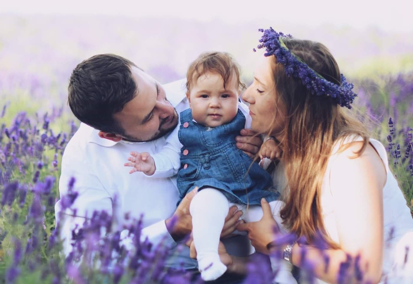 Rodzinne ujęcie w polu lawendy: tata trzyma uśmiechnięte dziecko w dżinsowej sukience, mama z wiankiem na głowie patrzy z czułością.