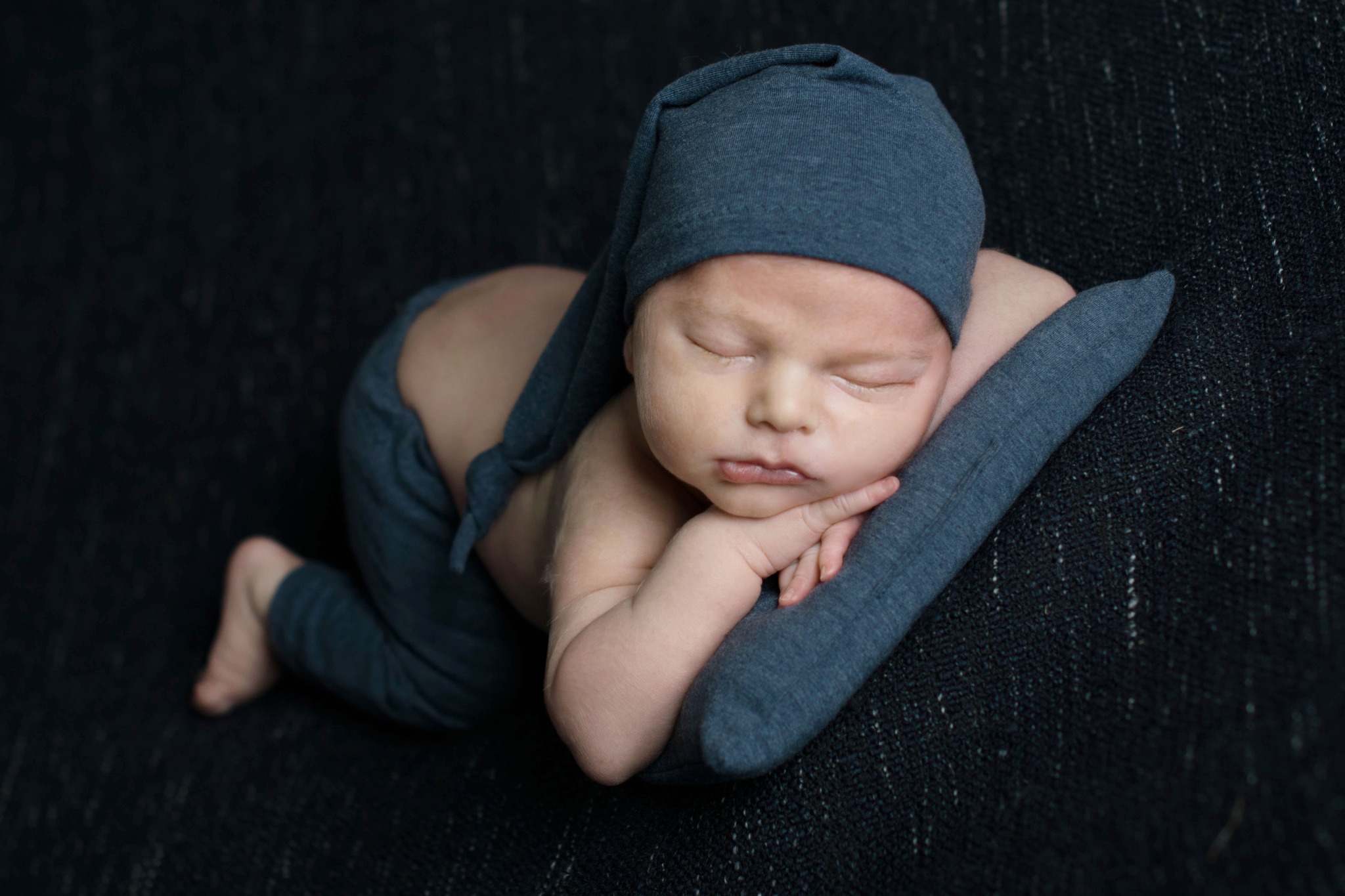 Śpiący noworodek w granatowym stroju i czapce, ułożony na poduszce na ciemnym tle.