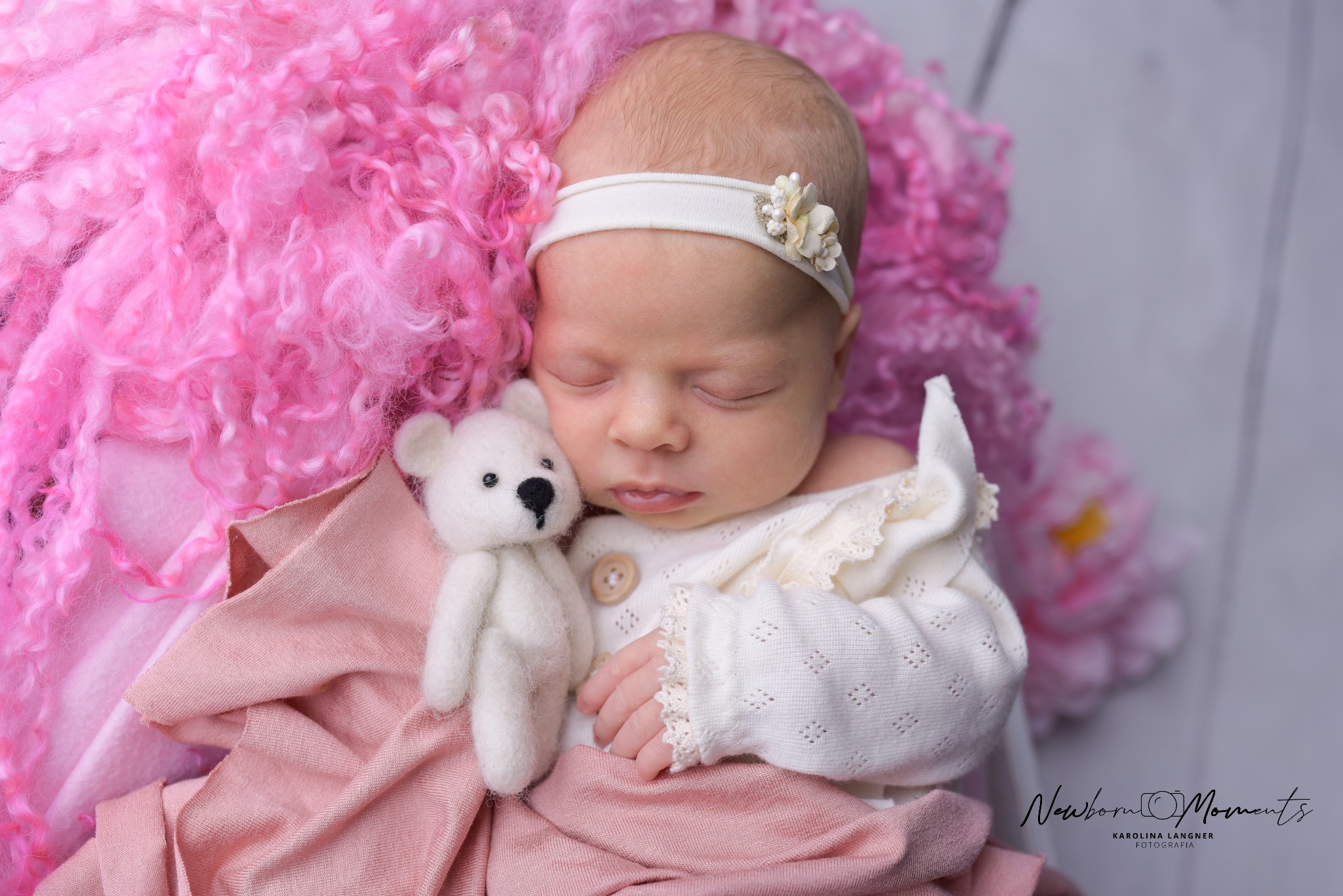 Śpiące nowonarodzone dziecko z opaską z kwiatem na głowie, trzymające pluszowego misia na tle różowych tekstur
