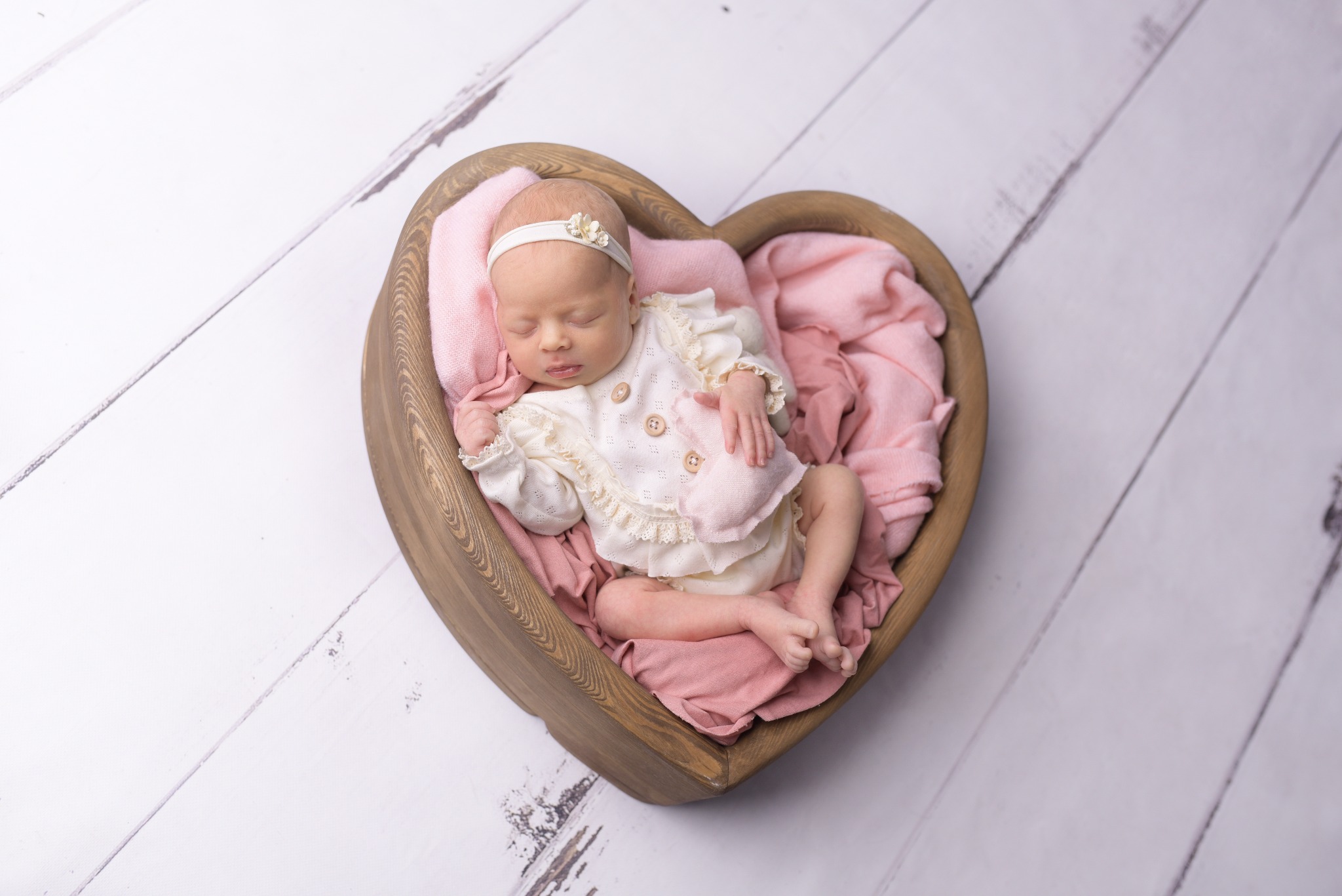 Śpiąca noworodka w drewnianej misce w kształcie serca, otulona różową tkaniną, ubrana w biały, koronkowy strój i opaskę z kwiatkiem, na tle białych desek.