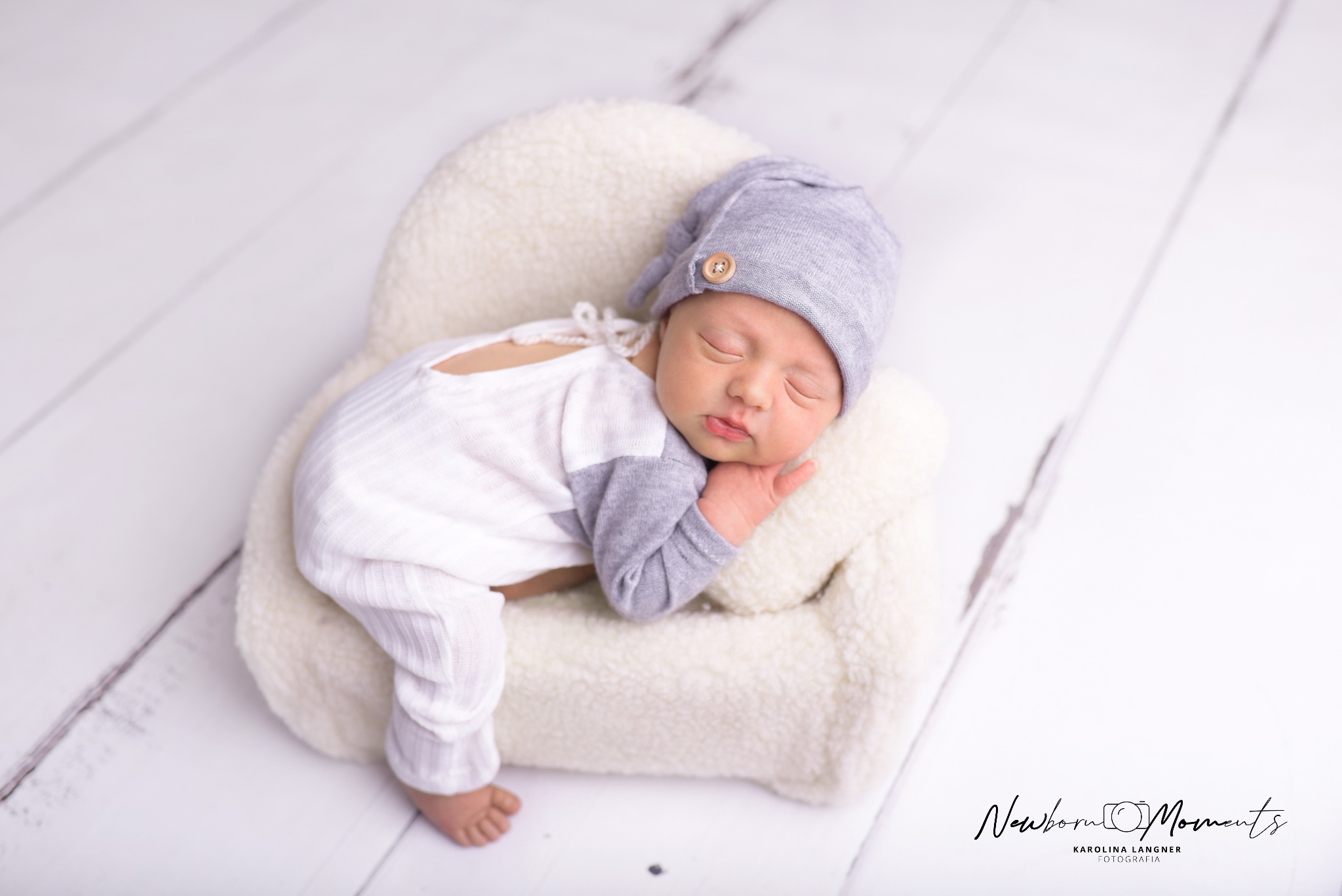 Śpiący noworodek w szaro-białym ubranku i czapeczce z guzikiem, ułożony na małym, pluszowym fotelu na tle białych desek.