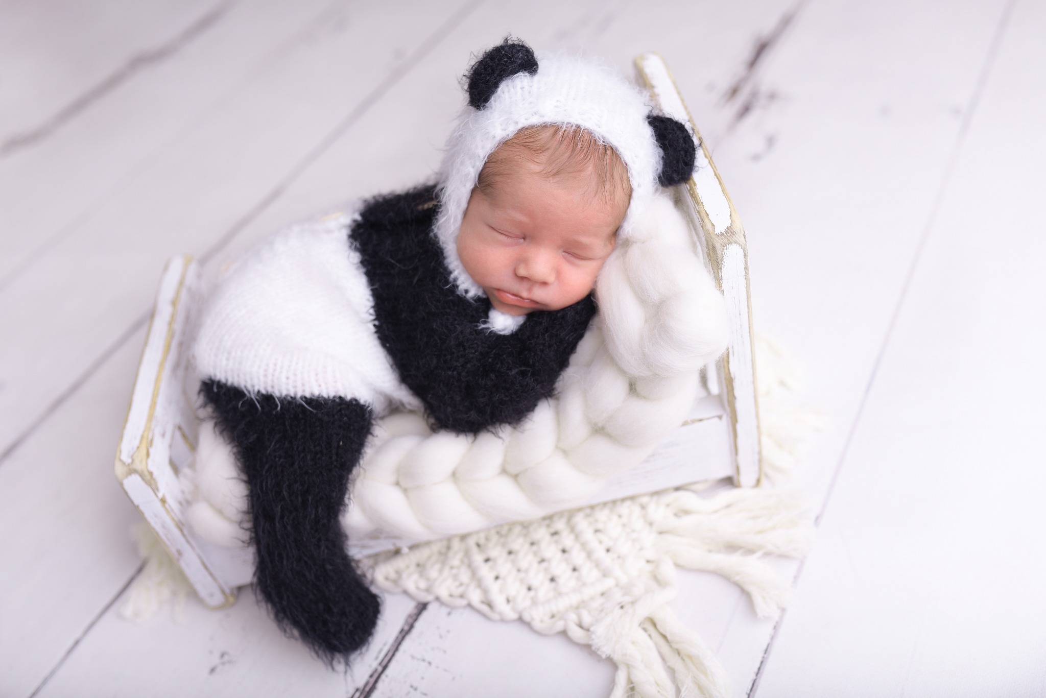 Śpiący noworodek w kostiumie pandy, ułożony na miniaturowym, białym łóżeczku z wełnianym materacem na tle białych drewnianych desek.