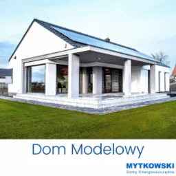 Nowoczesny dom jednorodzinny z białymi ścianami, płaskim dachem z panelami słonecznymi i dużymi oknami tarasowymi, otoczony zielonym trawnikiem.