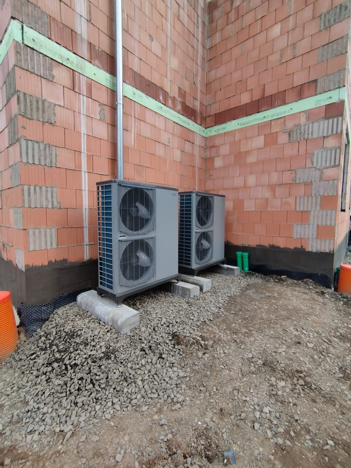 Dwie szare jednostki zewnętrzne pompy ciepła zamontowane na betonowych podstawach przed ścianą z czerwonej cegły w trakcie budowy, z widoczną rurą pionową i elementami instalacji.