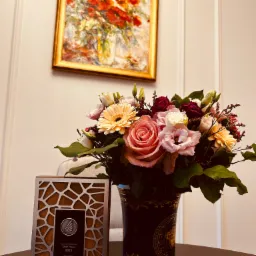 Bukiet różowych i kremowych kwiatów w ozdobnym wazonie Versace, obok nagroda 'Laureat Konkursu Orły Prawa 2023' na ciemnym stoliku, w tle obraz w złotej ramie.