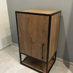 Szafka z drewnopodobną okleiną i czarną metalową ramą, z półką u dołu, na tle szarej ściany i marmurowej podłogi.