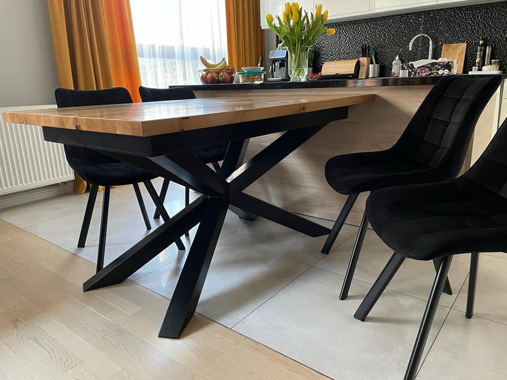 Rozkładany stół z drewnianym blatem i czarną, metalową podstawą w kształcie litery X, otoczony czarnymi krzesłami w nowoczesnej kuchni z żółtymi tulipanami w wazonie i owocami w tle.