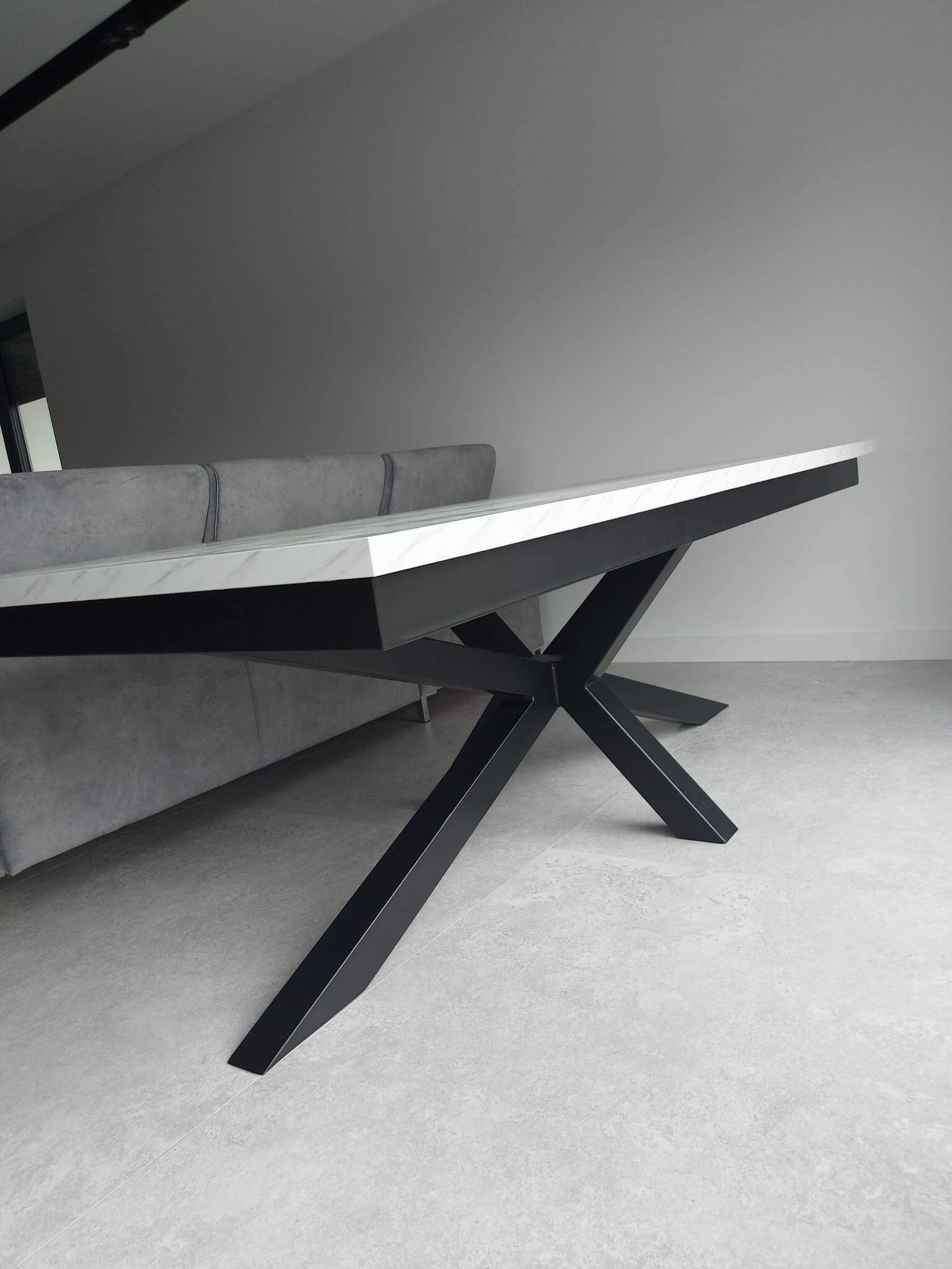 Stół z białym blatem imitującym marmur i czarną, geometryczną podstawą z metalu, widoczny fragment szarej sofy w tle.