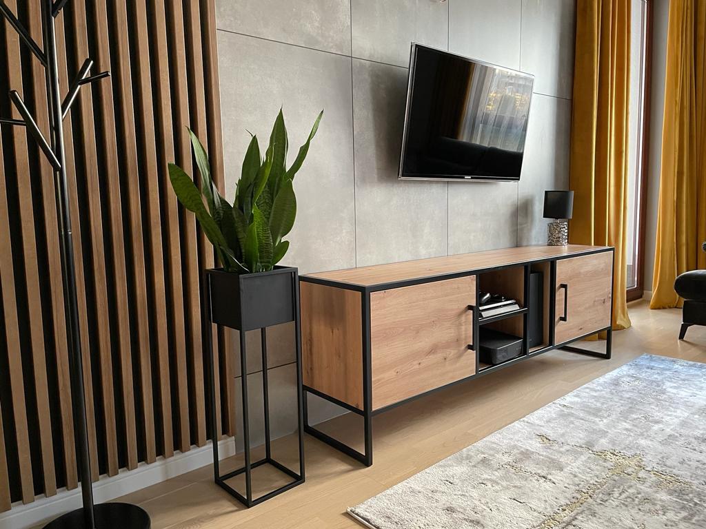Nowoczesna szafka RTV z drewnianymi frontami i czarnymi metalowymi nogami, podwieszony telewizor, dekoracyjne panele ścienne i roślina w metalowej donicy na tle żółtych zasłon.