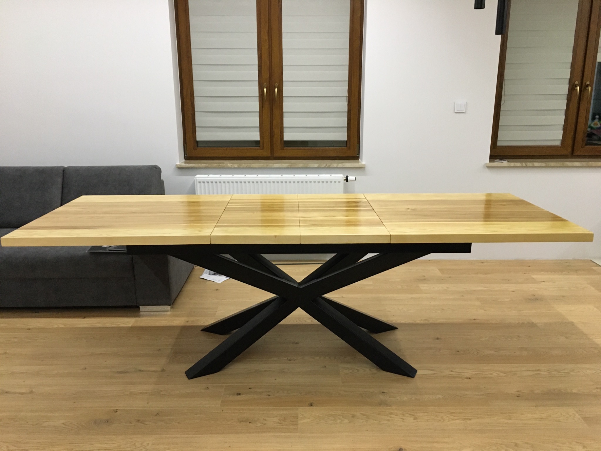 Rozkładany stół z jasnego drewna z czarną, geometryczną podstawą w kształcie litery X, ustawiony na drewnianej podłodze w jasnym wnętrzu.